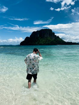 A man walks through clear tropical waters towards a lush island under a bright summer sky.
