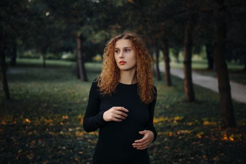 Free Portrait of a redheaded woman in a dark dress, standing amidst trees in a serene forest park during twilight. Stock Photo