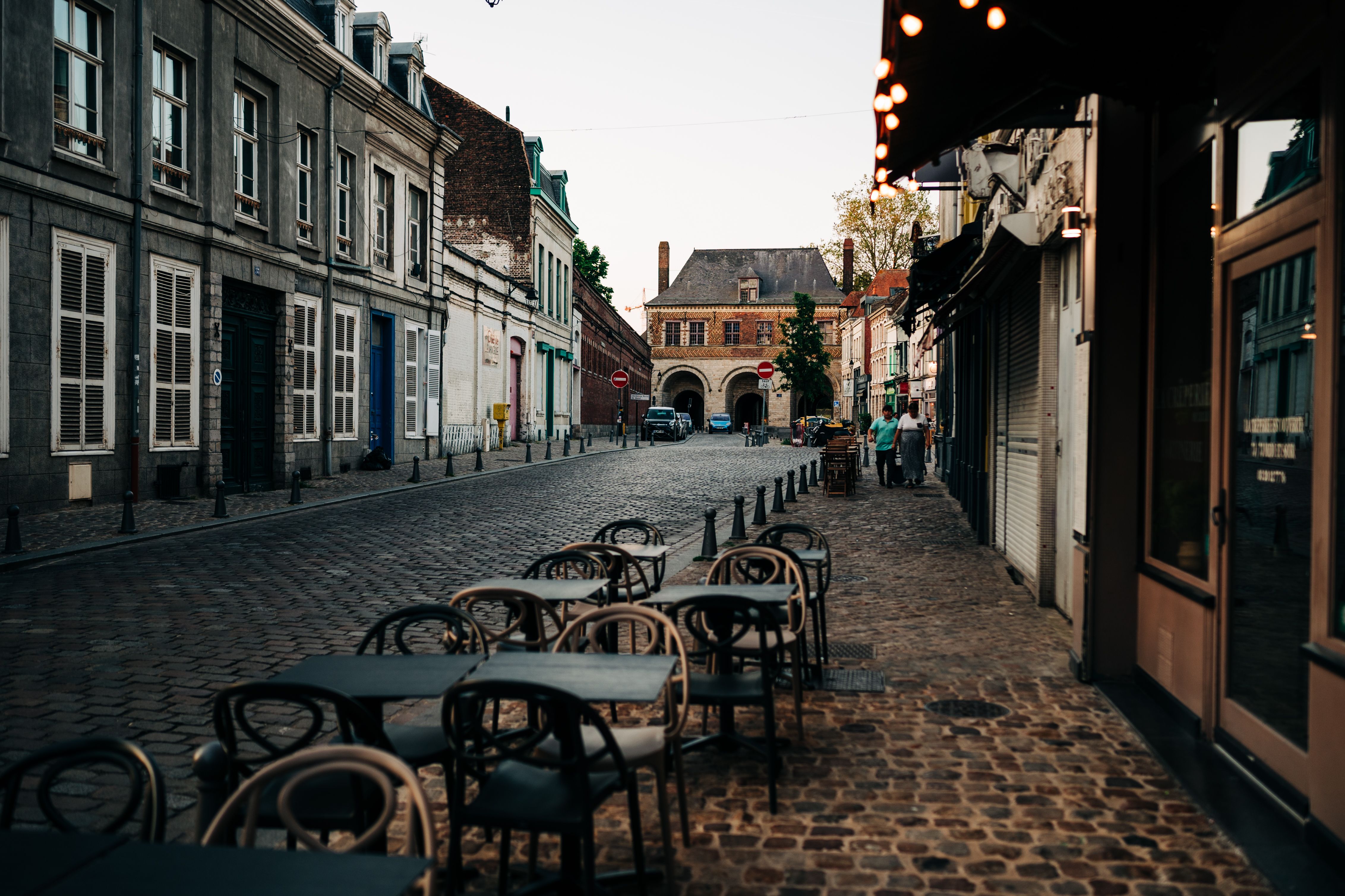 Outdoor café tables line a quiet cobbled street flanked by historic buildings, with an archway in the distance