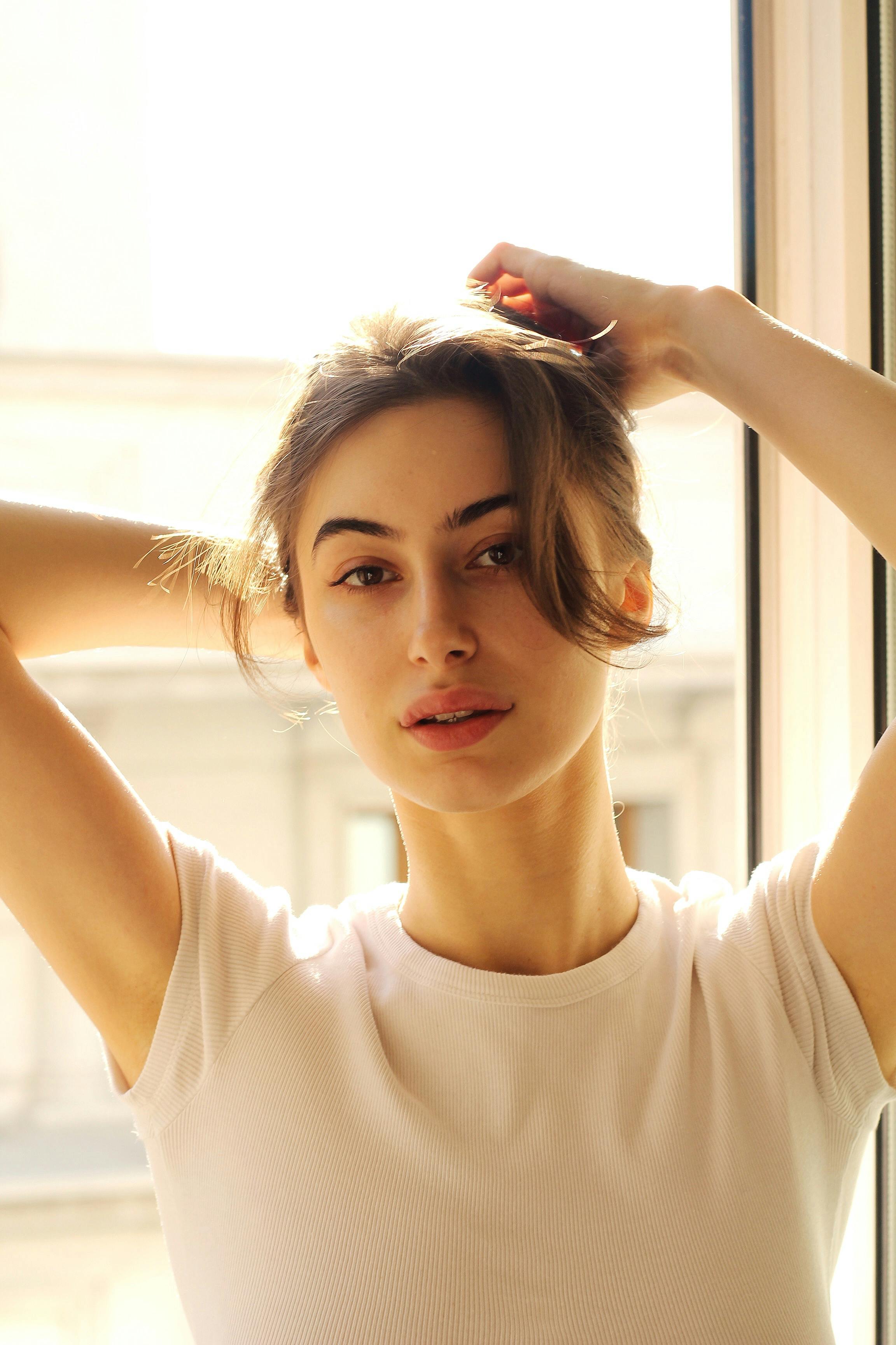 A young woman poses by a window with sunlight illuminating her face, creating a soft and natural look.