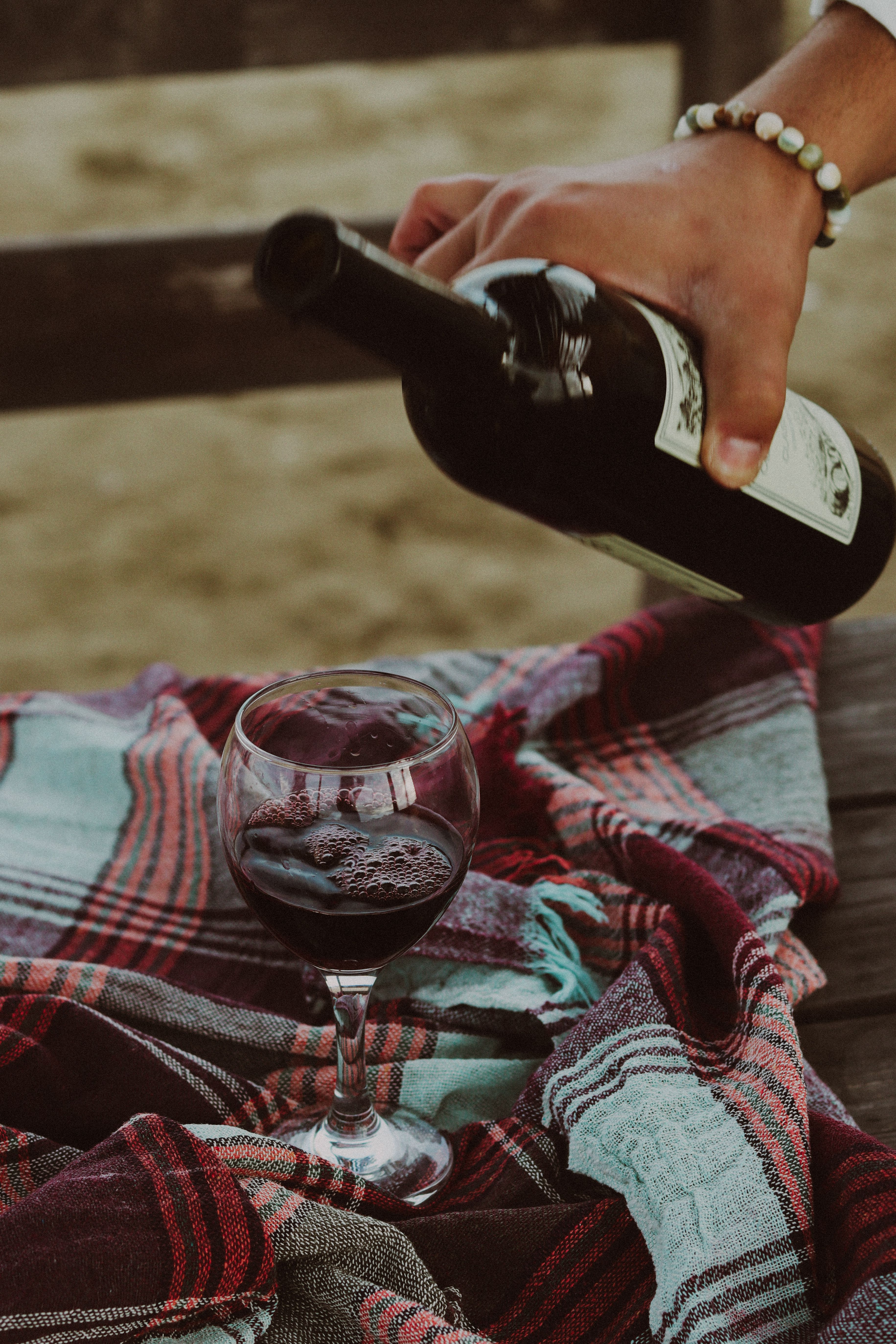 Close-up of hand pouring red wine into a glass on a plaid blanket during a picnic.