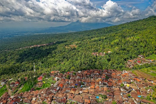 Aerial view of Majasari village with lush green forests and distant mountains under a cloudy sky.