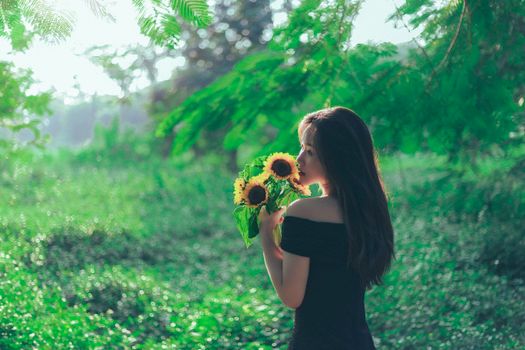 A young woman in a dark dress holds sunflowers in a vibrant, sunlit park setting.