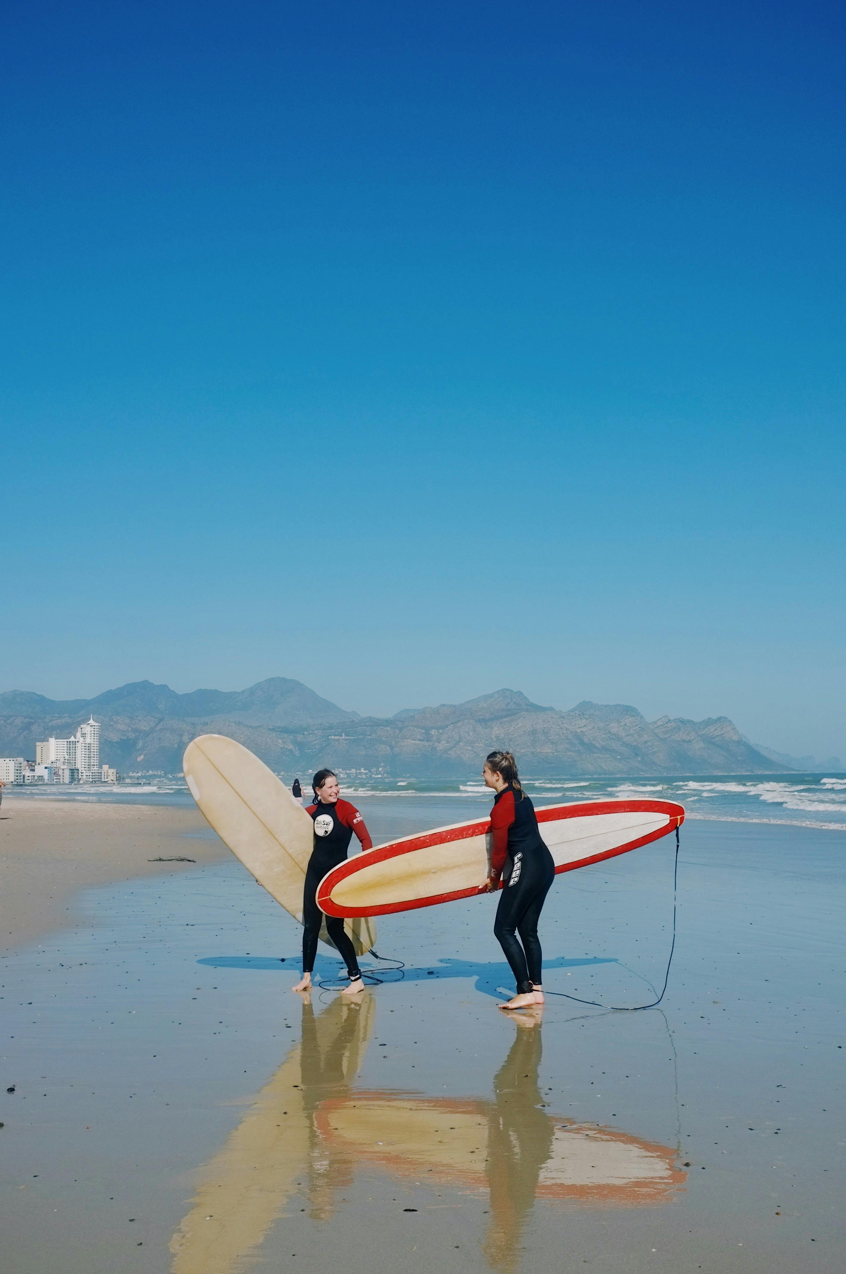 Two surfers with boards walking on a sunny beach with mountains in the background.