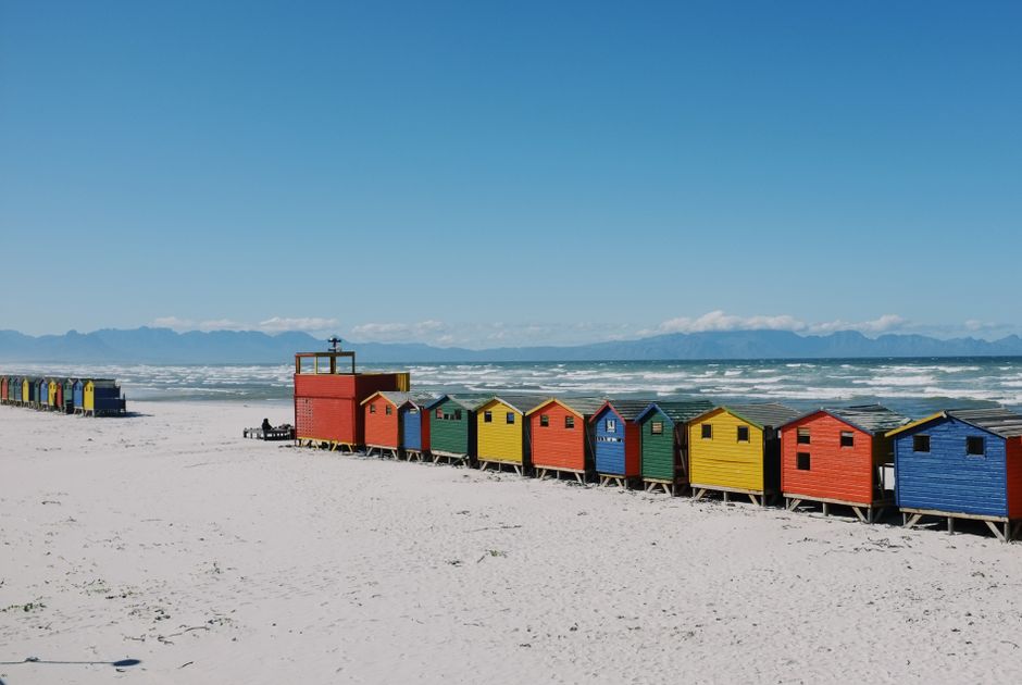 Colorful beach huts on a sandy shore with ocean waves and mountains in the background.