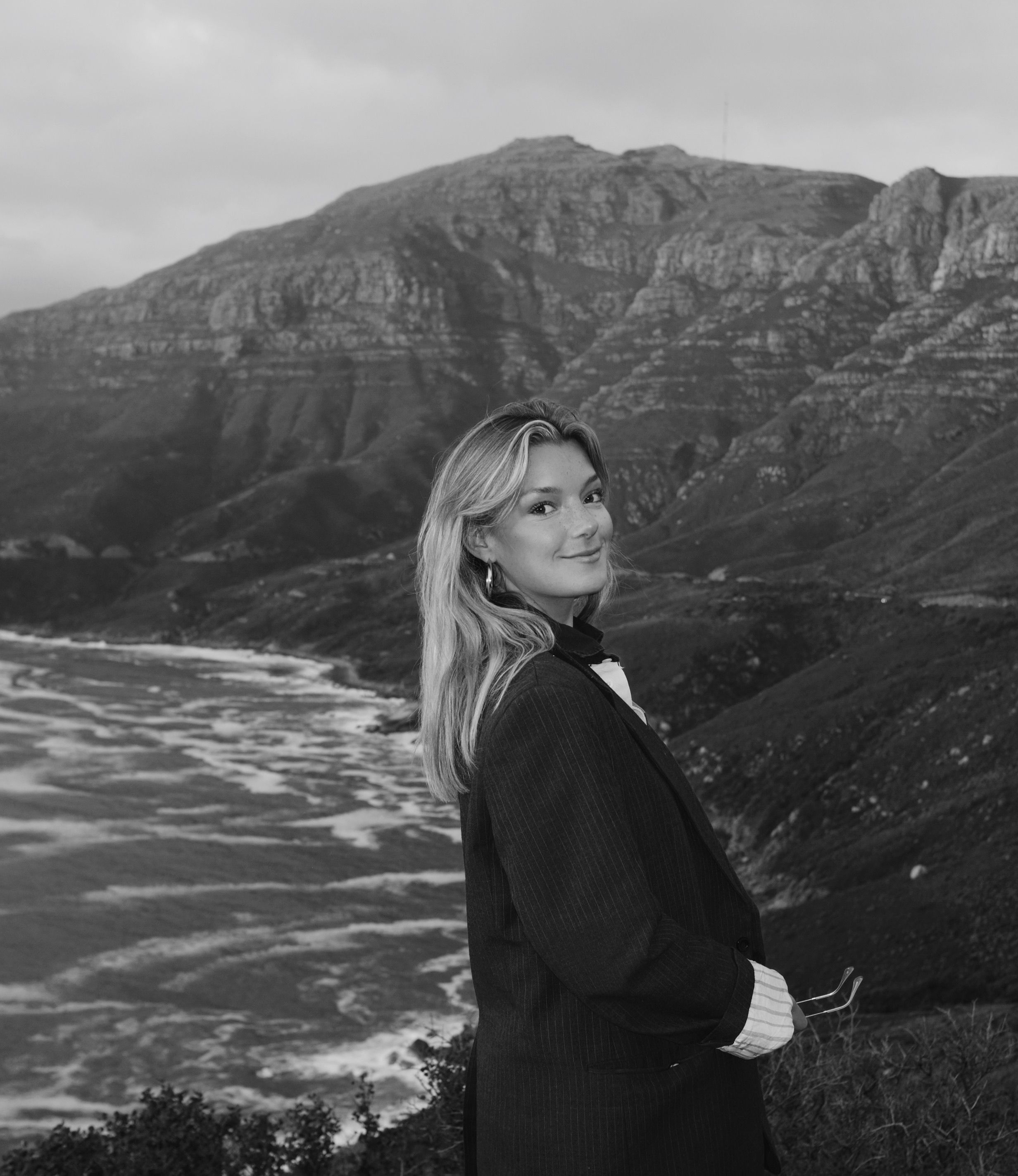 Black and white photo of a woman by mountains and seashore in a blazer.