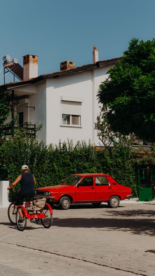 Free An elderly man rides a tricycle beside a vintage red car in a sunny Turkish neighborhood. Stock Photo