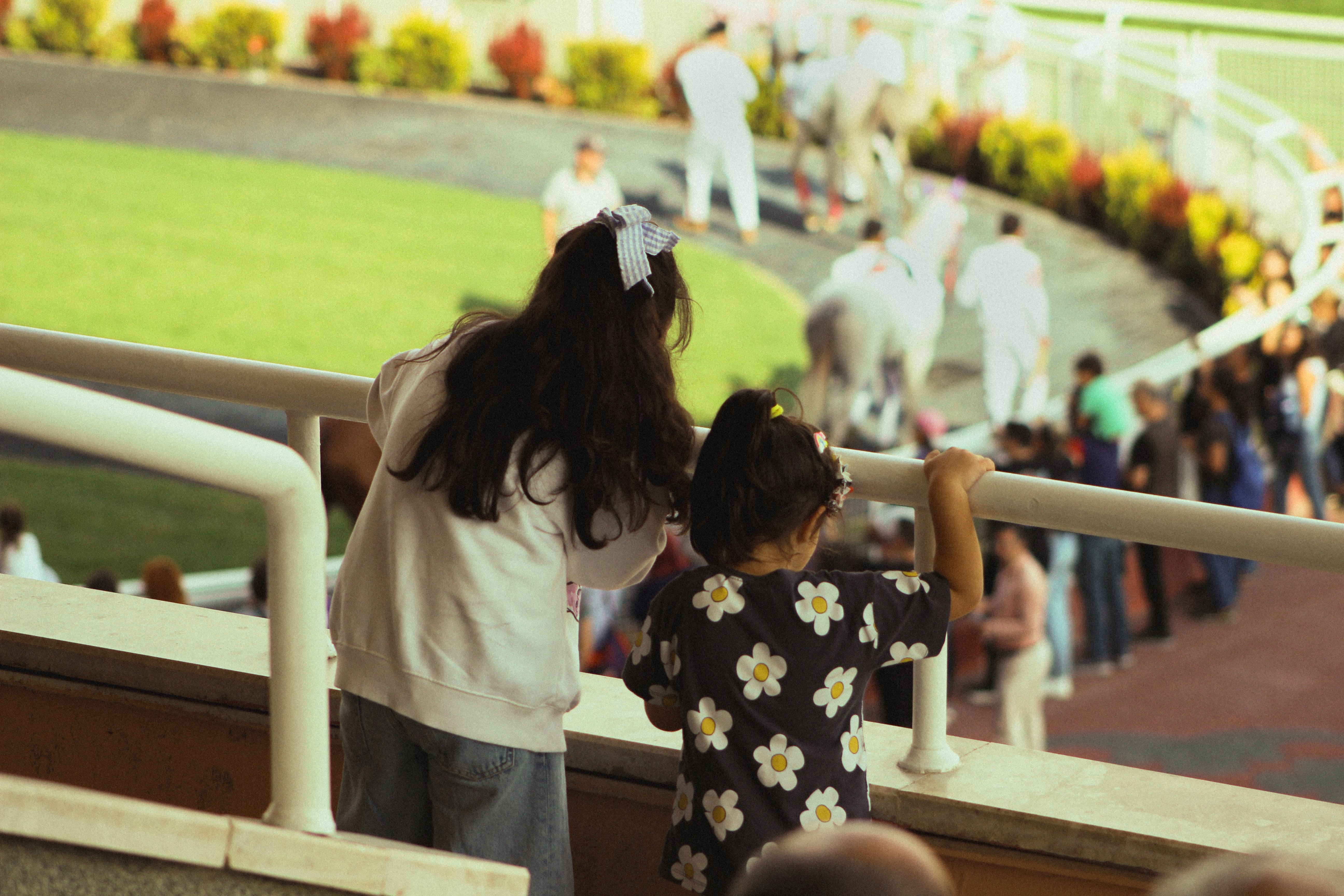 Two young girls lean over railings to watch a horse race, surrounded by a lively crowd. Outdoor recreational scene.