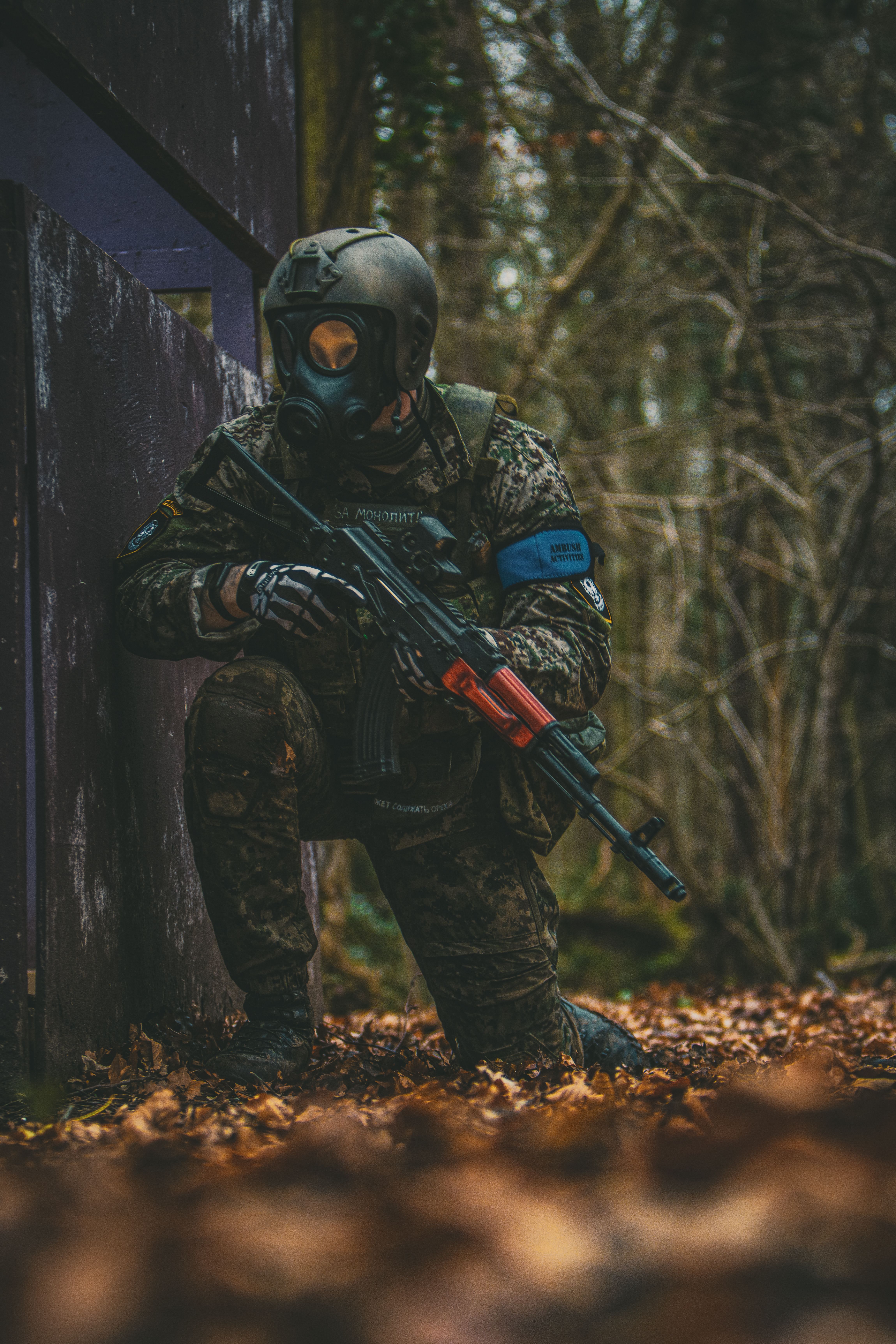 A soldier in camouflage kneels in a forest, wearing a gas mask and holding a rifle.