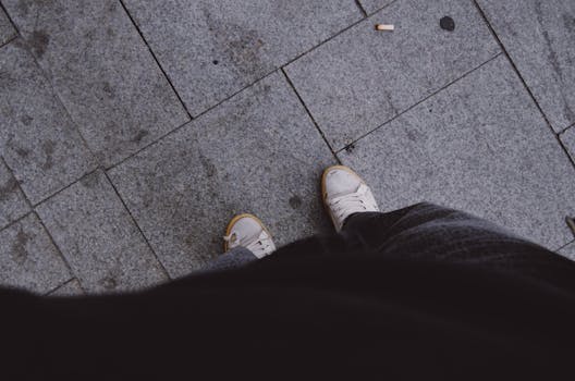 Overhead view of a person in sneakers on a paved street with a cigarette butt nearby.