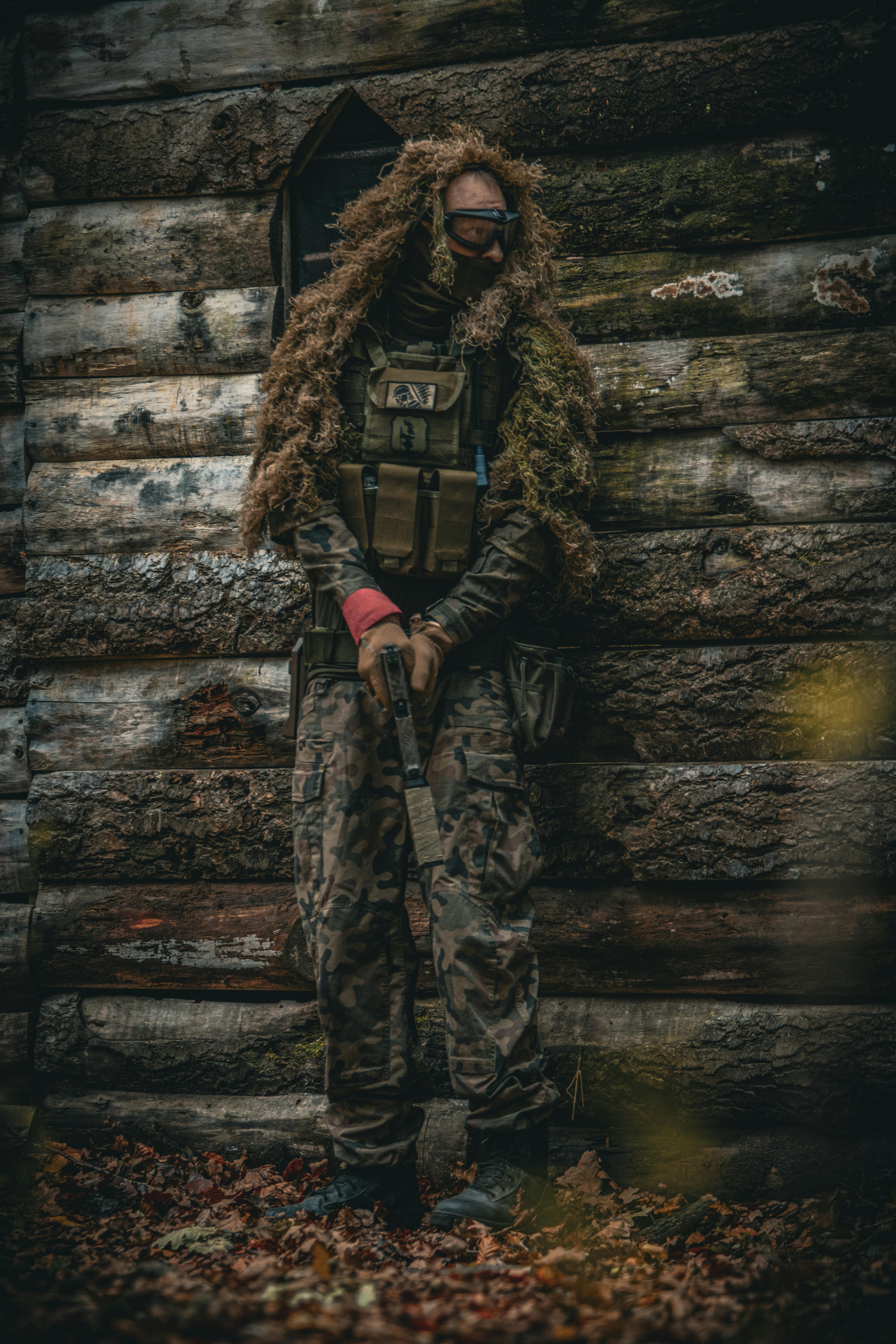 Eine Person in Tarnkleidung mit einem Paintballgewehr steht im Freien an der Wand einer Blockhütte. Prescription Goggle Inserts - A person in camouflage gear holding a paintball gun stands by a log cabin wall outdoors.