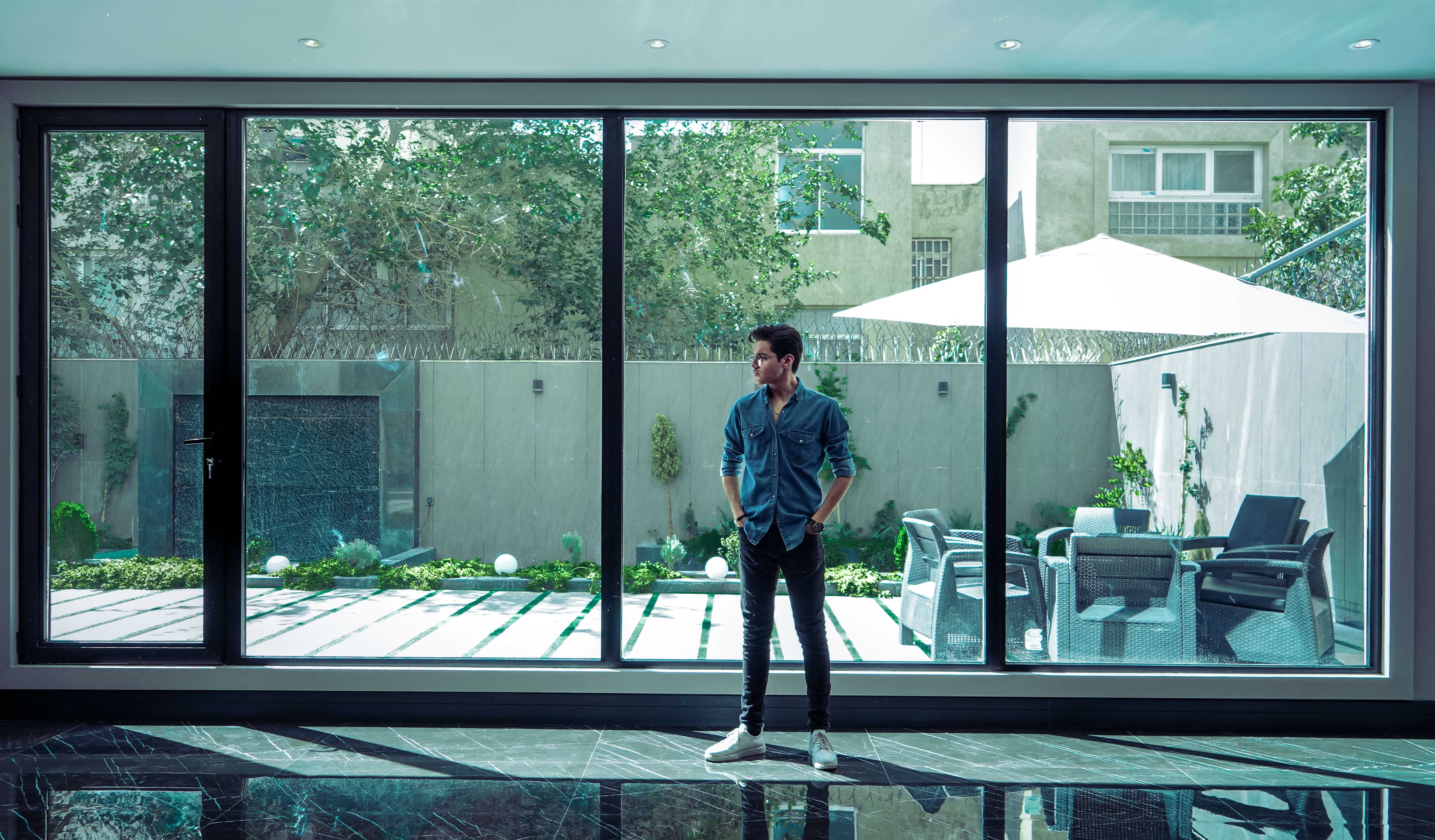 Stylish young man standing in a modern room with large glass windows overlooking a garden.