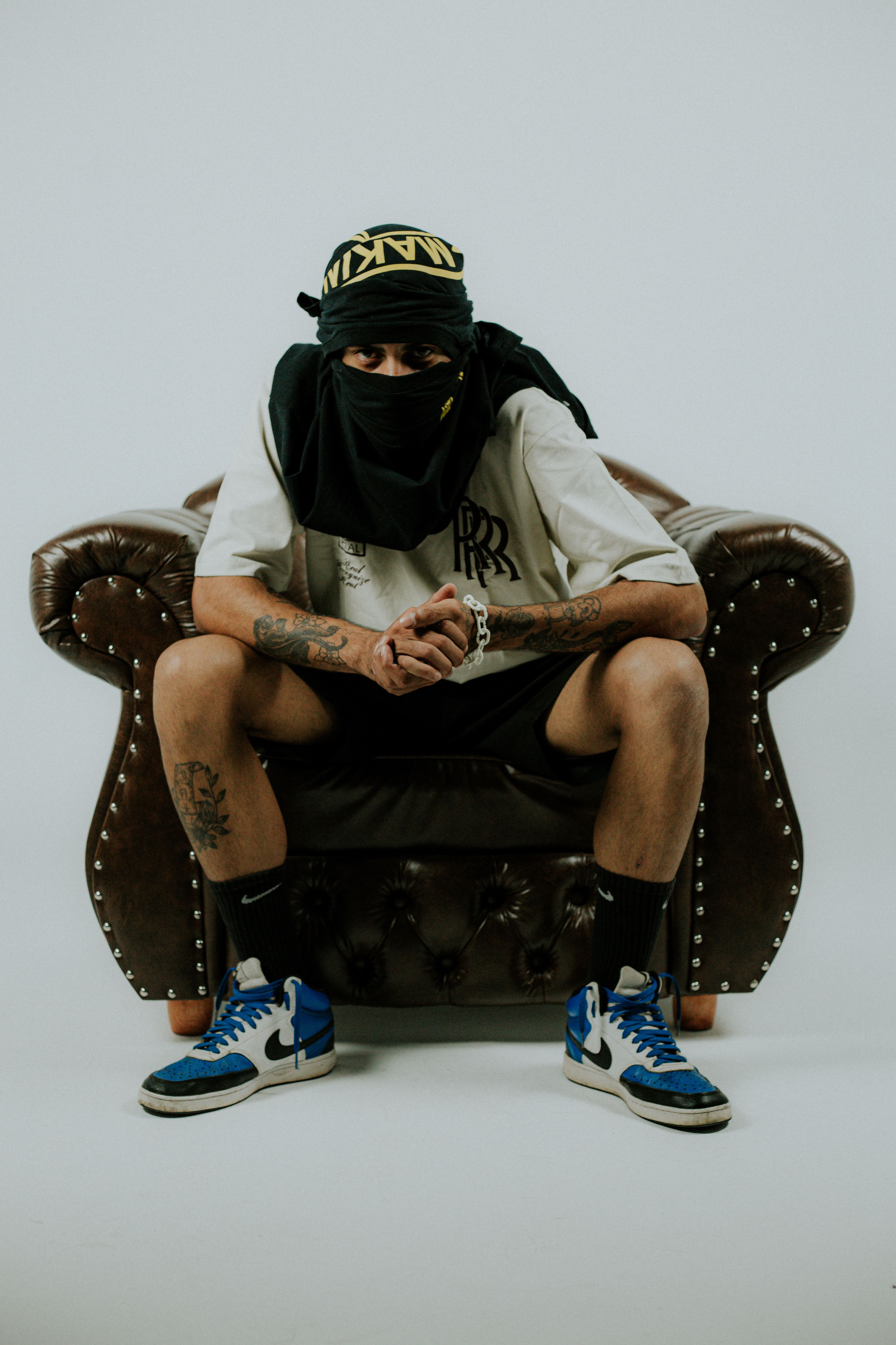 Man in streetwear sits on a leather armchair, wearing a balaclava and tattoos, against a white background.
