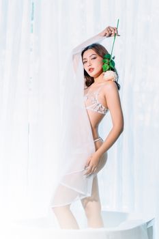 A woman posing in lingerie with a white rose in a bright setting.