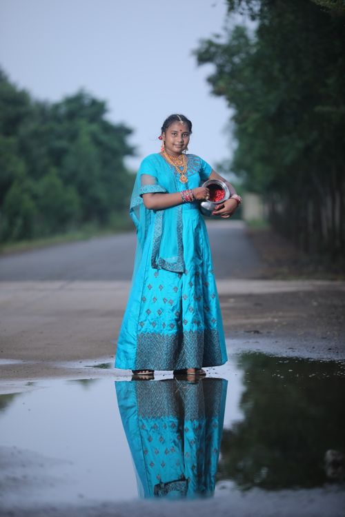 Free A woman in a blue traditional dress holding a pot stands by a puddle reflecting her image on a rural road. Stock Photo