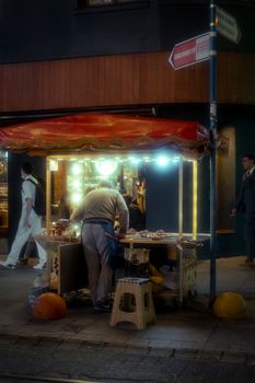 A lively street market scene at night with a vendor stall illuminated by neon lights.