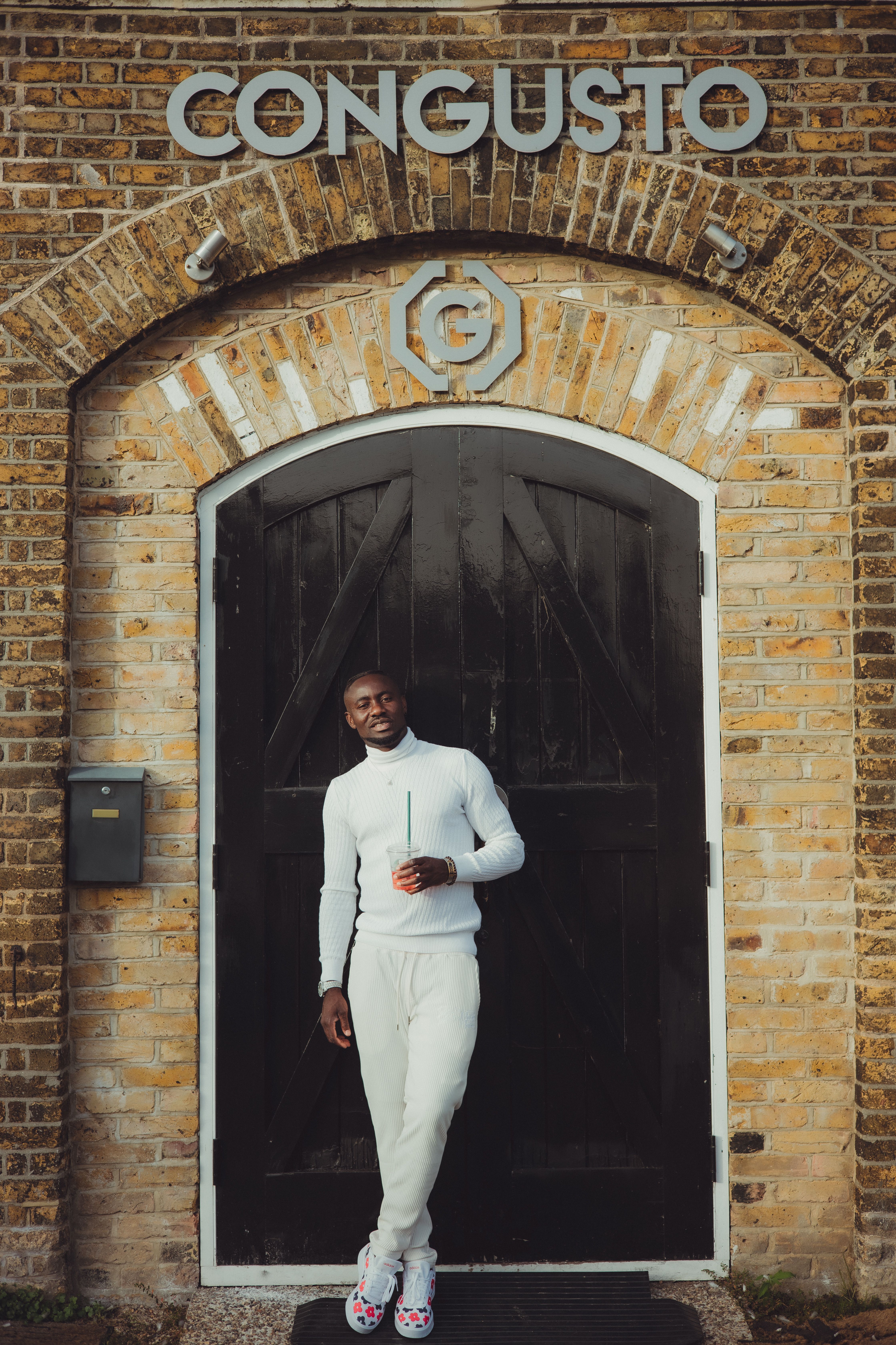 Fashionable man in white outfit holding cup, posing at rustic brick archway with large black door