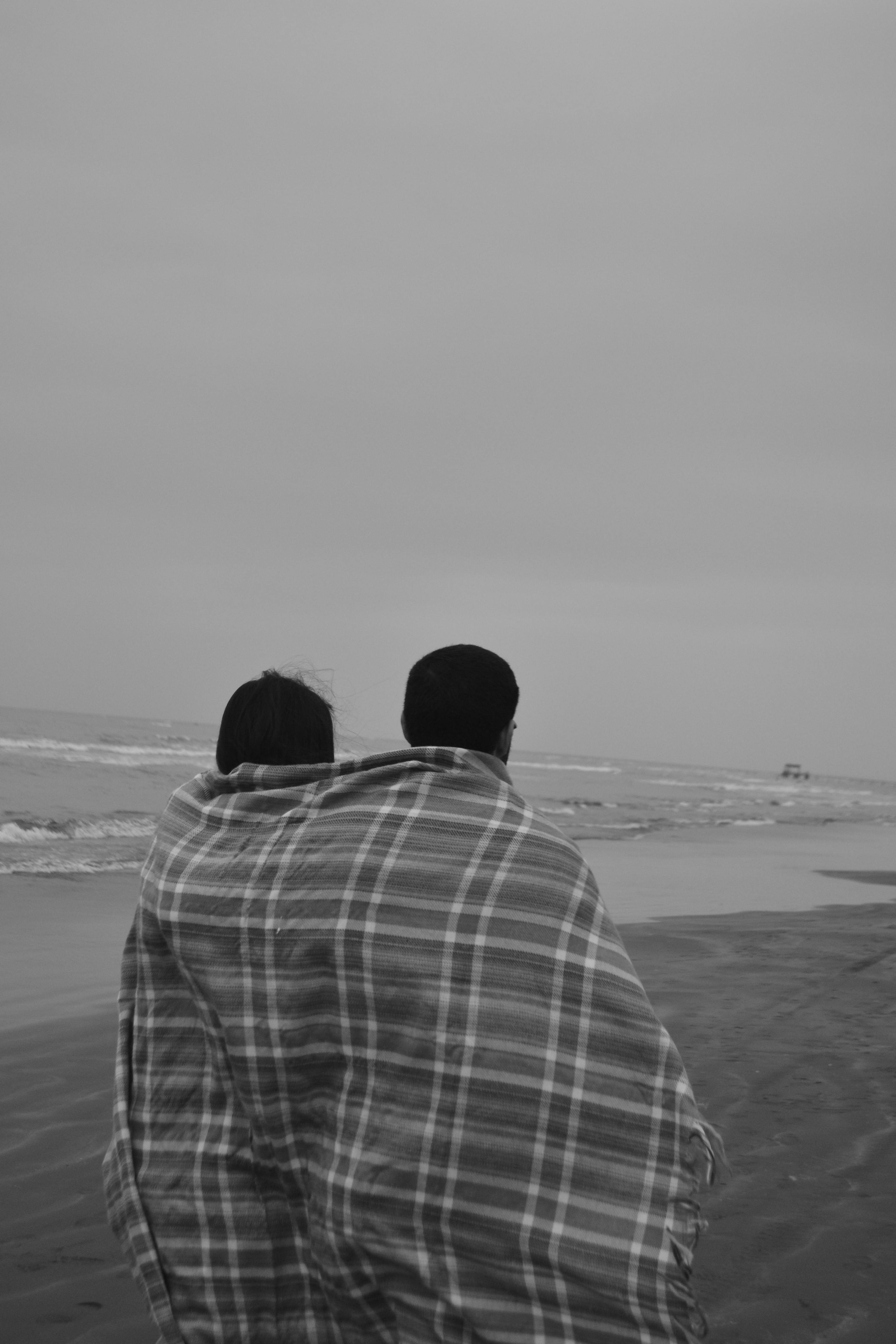 Black and white image of a couple embracing under a blanket on a windswept beach.