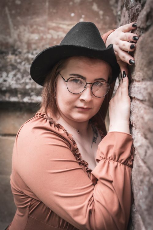 Free Portrait of a woman wearing glasses and a hat, posing by a rustic stone wall in Charfield, England. Stock Photo