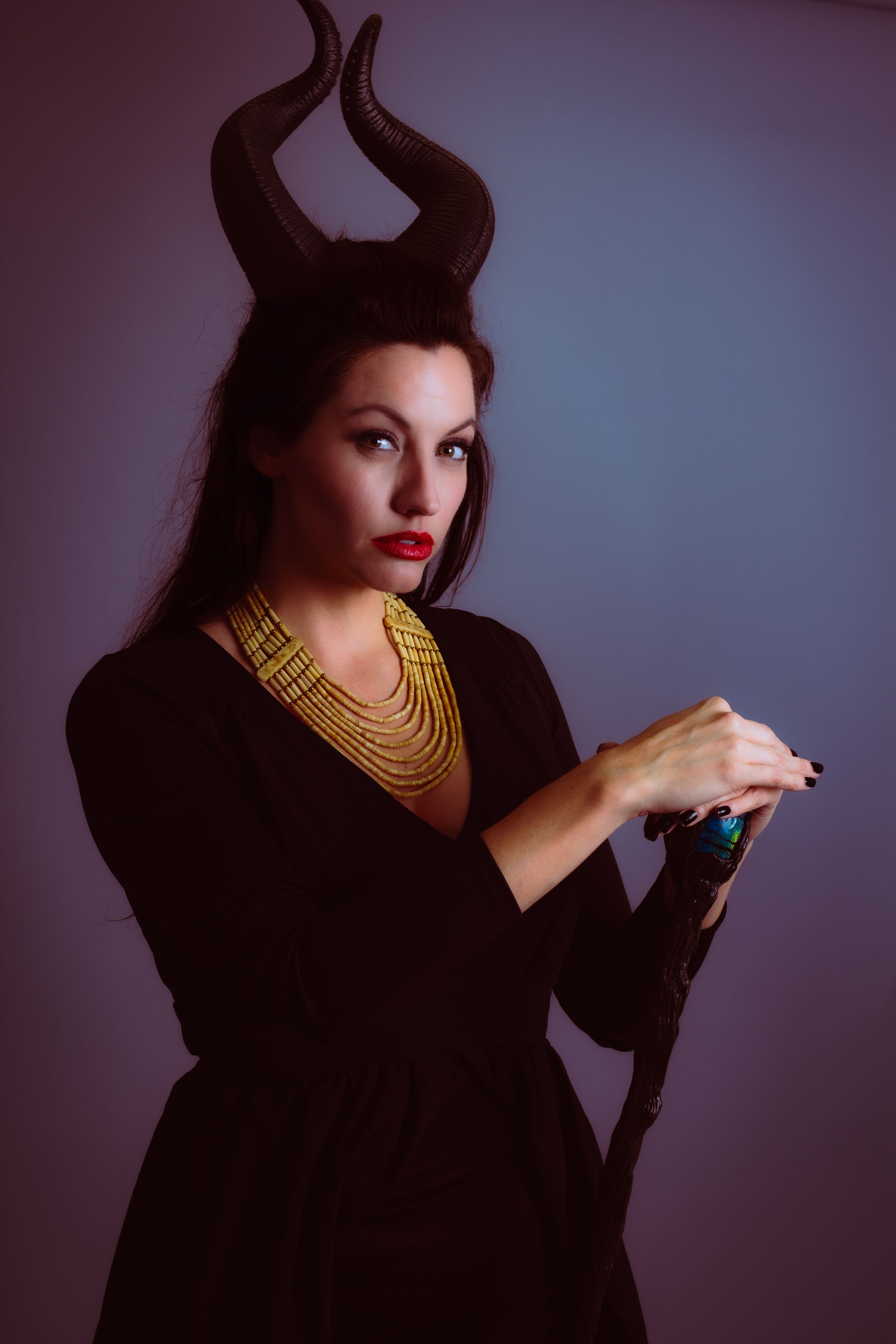 A woman in a black dress with horns and red lips poses with a staff. Studio shoot.