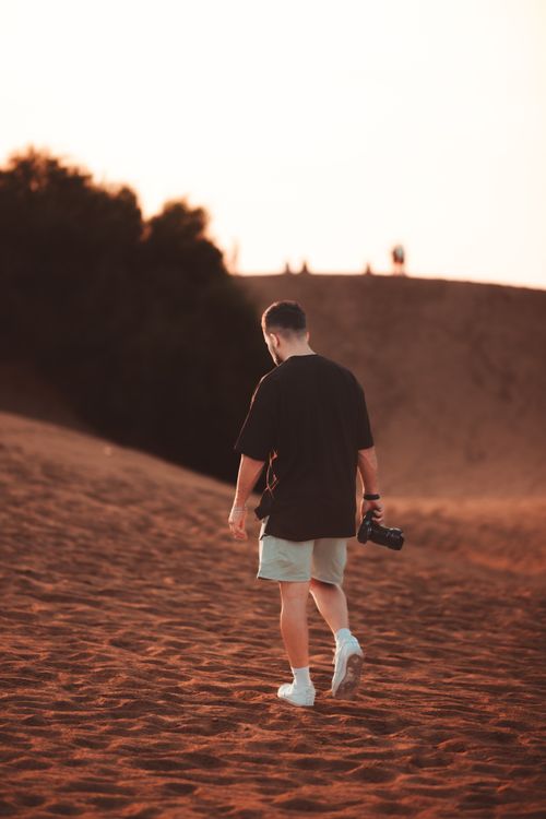 Free A man with a camera walks across sandy desert dunes during sunset. Stock Photo