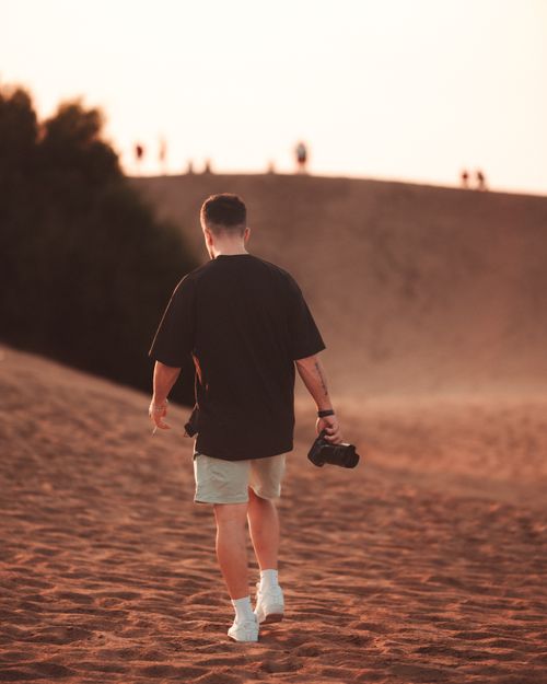 Free Silhouette of a man carrying a camera, strolling through desert dunes during a warm sunset. Stock Photo