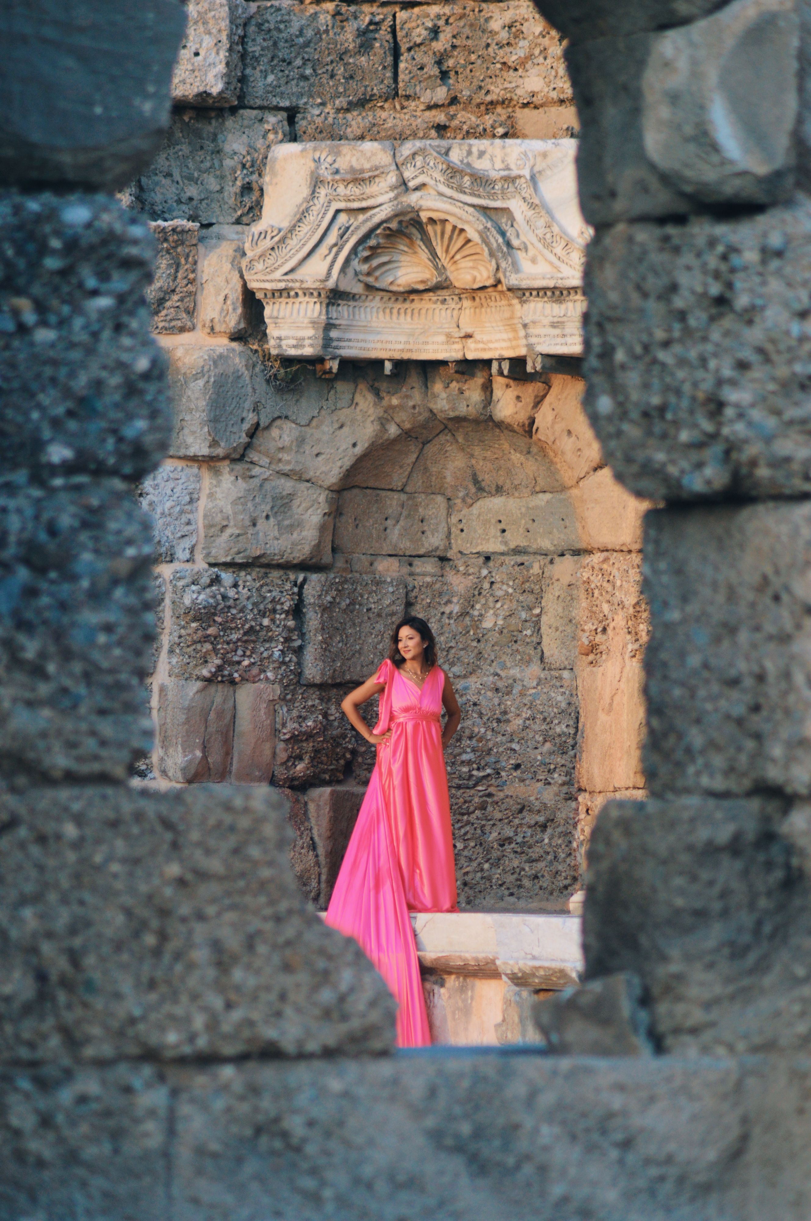 A woman in a vibrant pink dress poses gracefully against historical stone ruins.