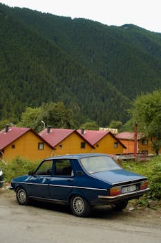 A blue vintage car parked in a quaint village with colorful houses and lush hills in the background.