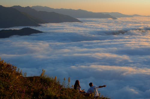 Woman and Man Sitting on Hill over Clouds at Sunset