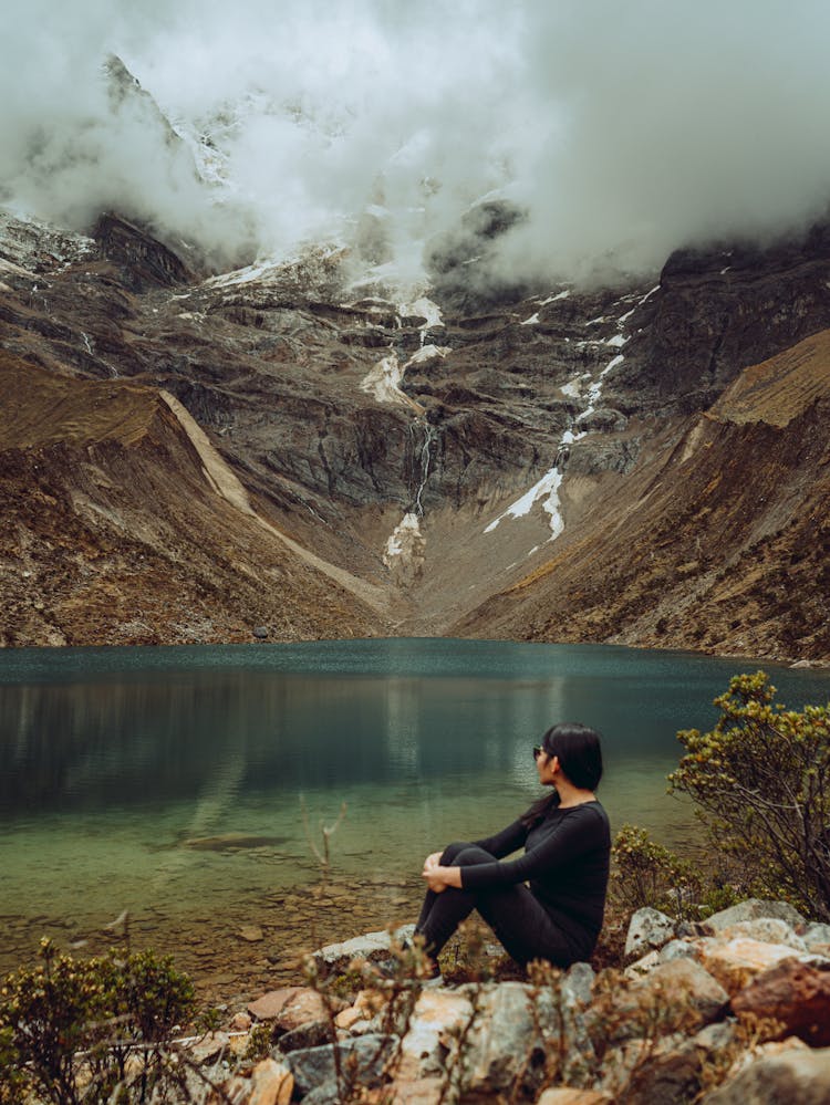 Woman Sits By Lake In Mountains