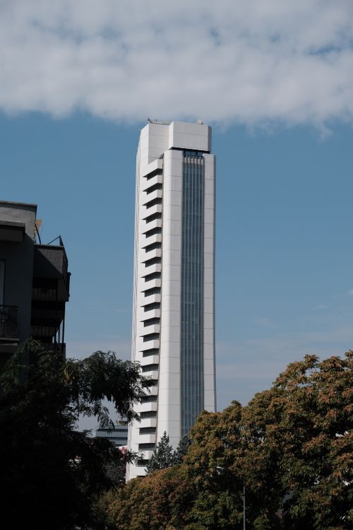 Free A sleek modern skyscraper stands tall against a clear blue sky with trees in the foreground. Stock Photo