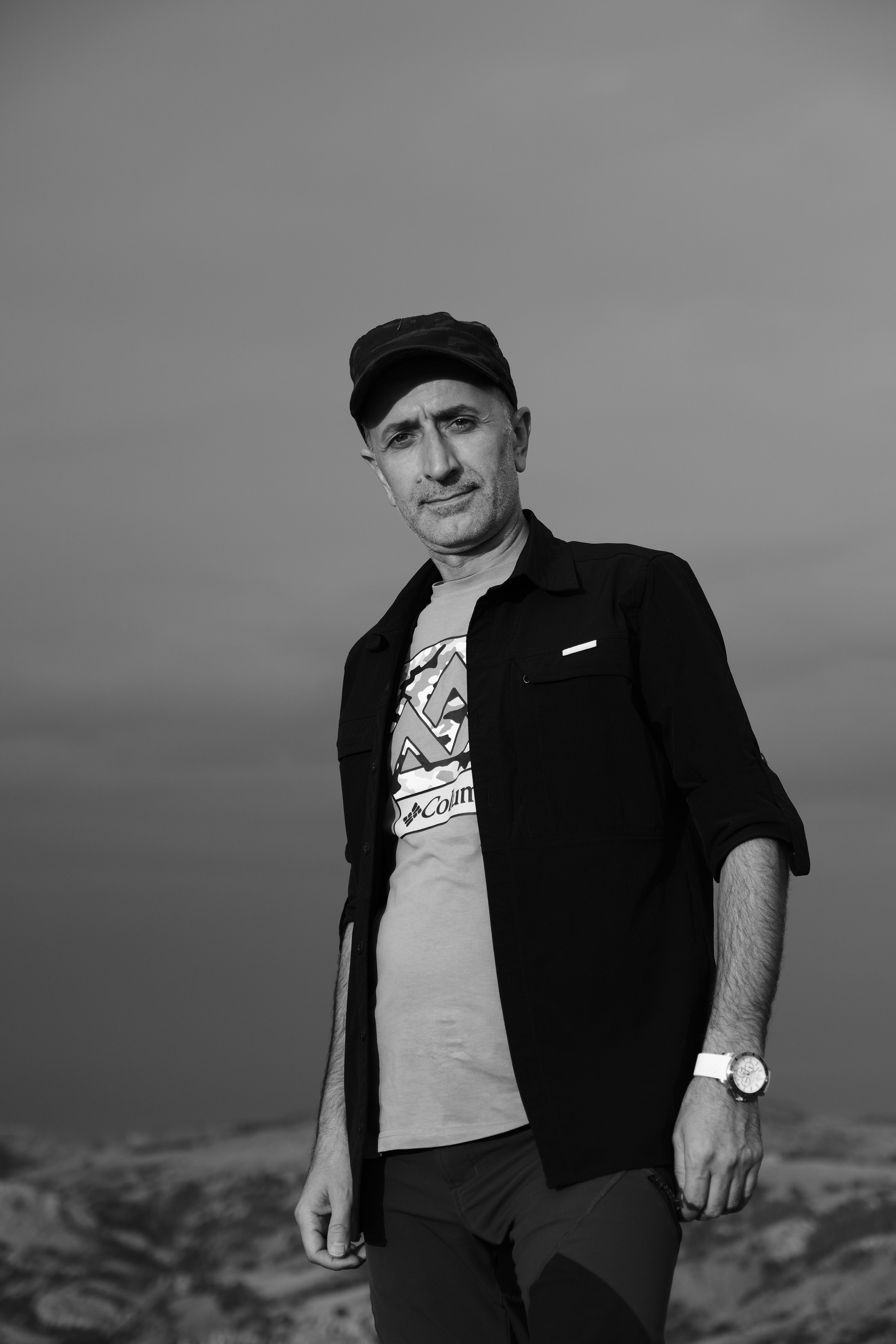 Black and white portrait of a man wearing a cap and black shirt in an outdoor setting.