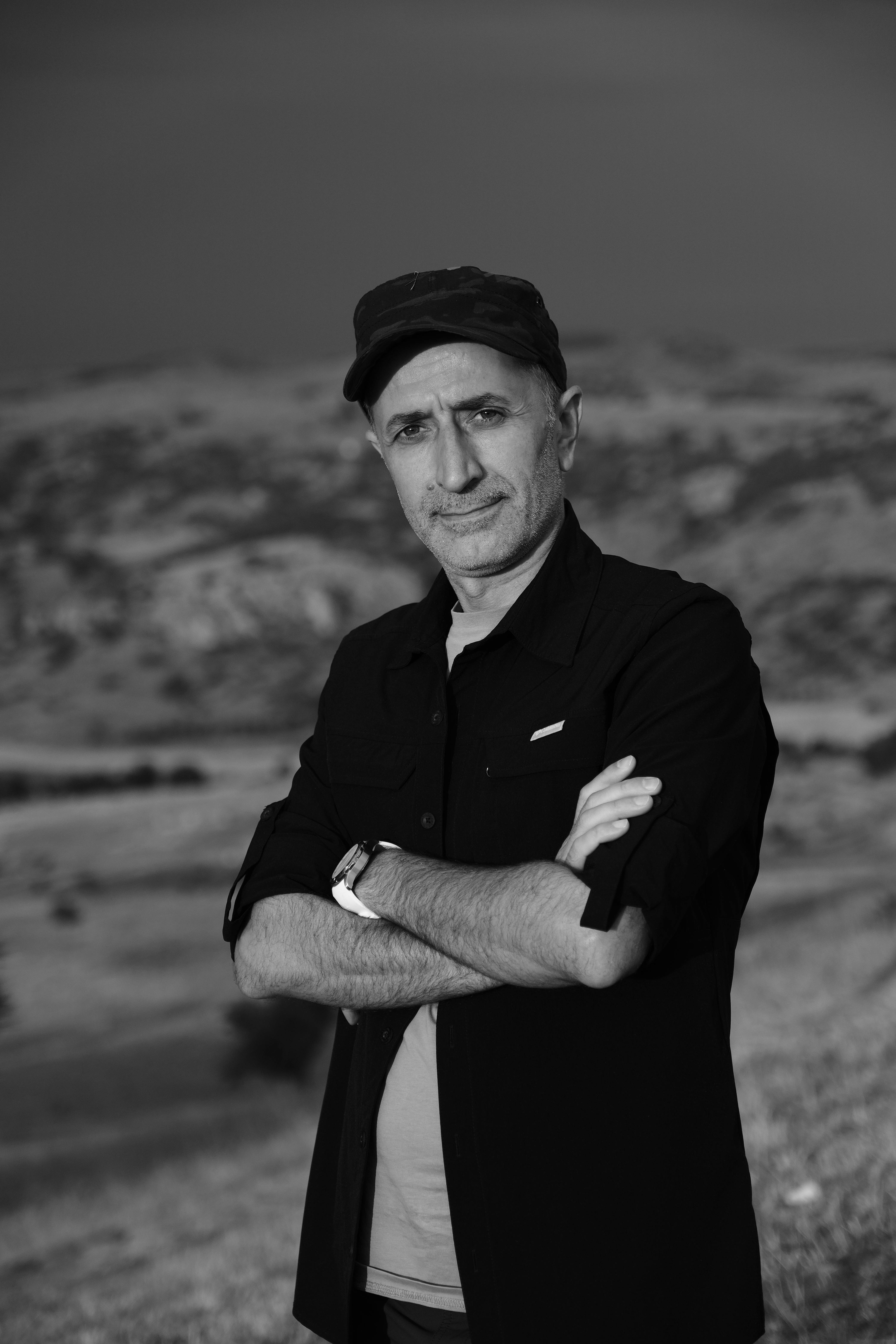 A monochrome portrait of a man in a cap with crossed arms, set in a scenic outdoor landscape.