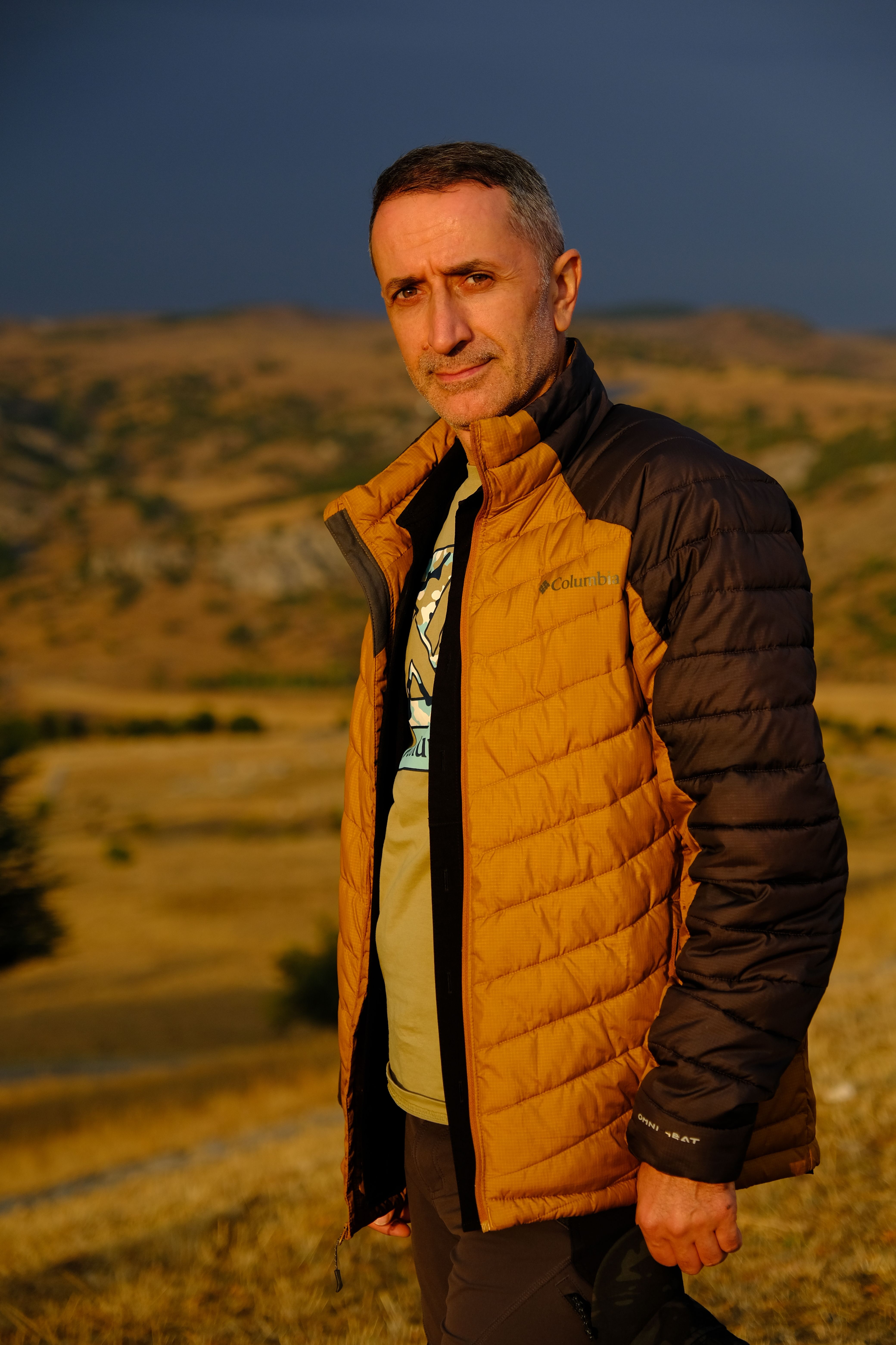 A man stands outdoors in a warm jacket during autumn, with scenic hills in the background.