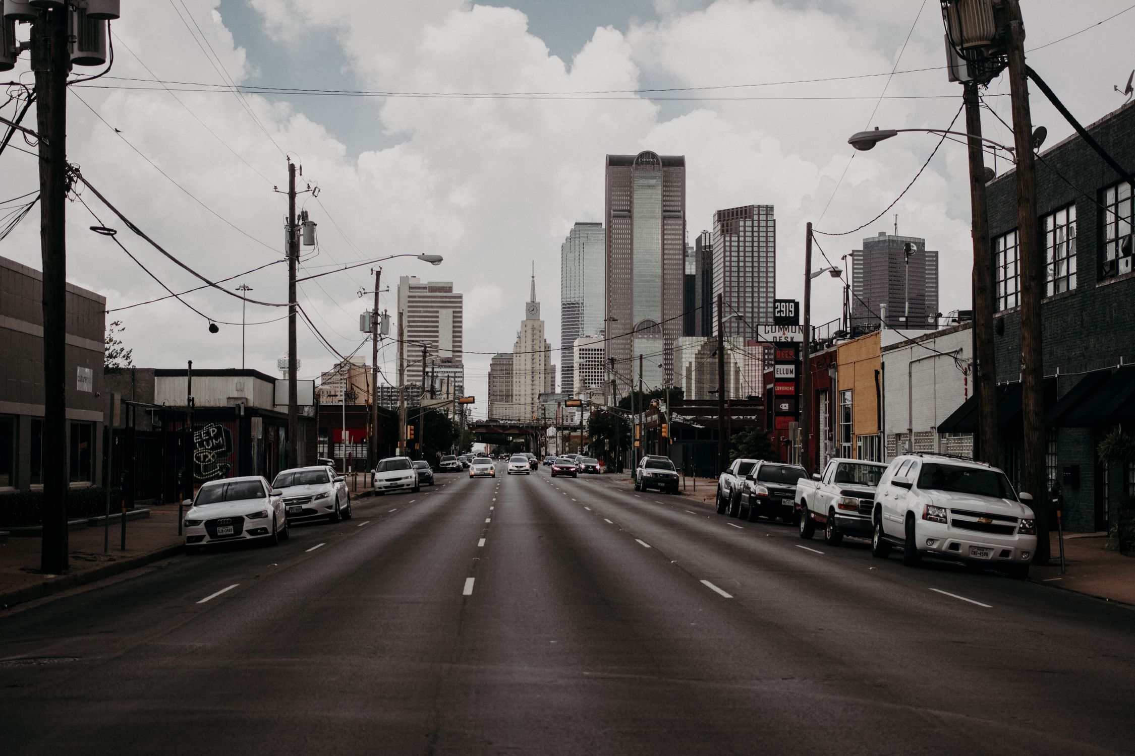 A city street leads to towering skyscrapers under an overcast sky, featuring cars and urban infrastructure.