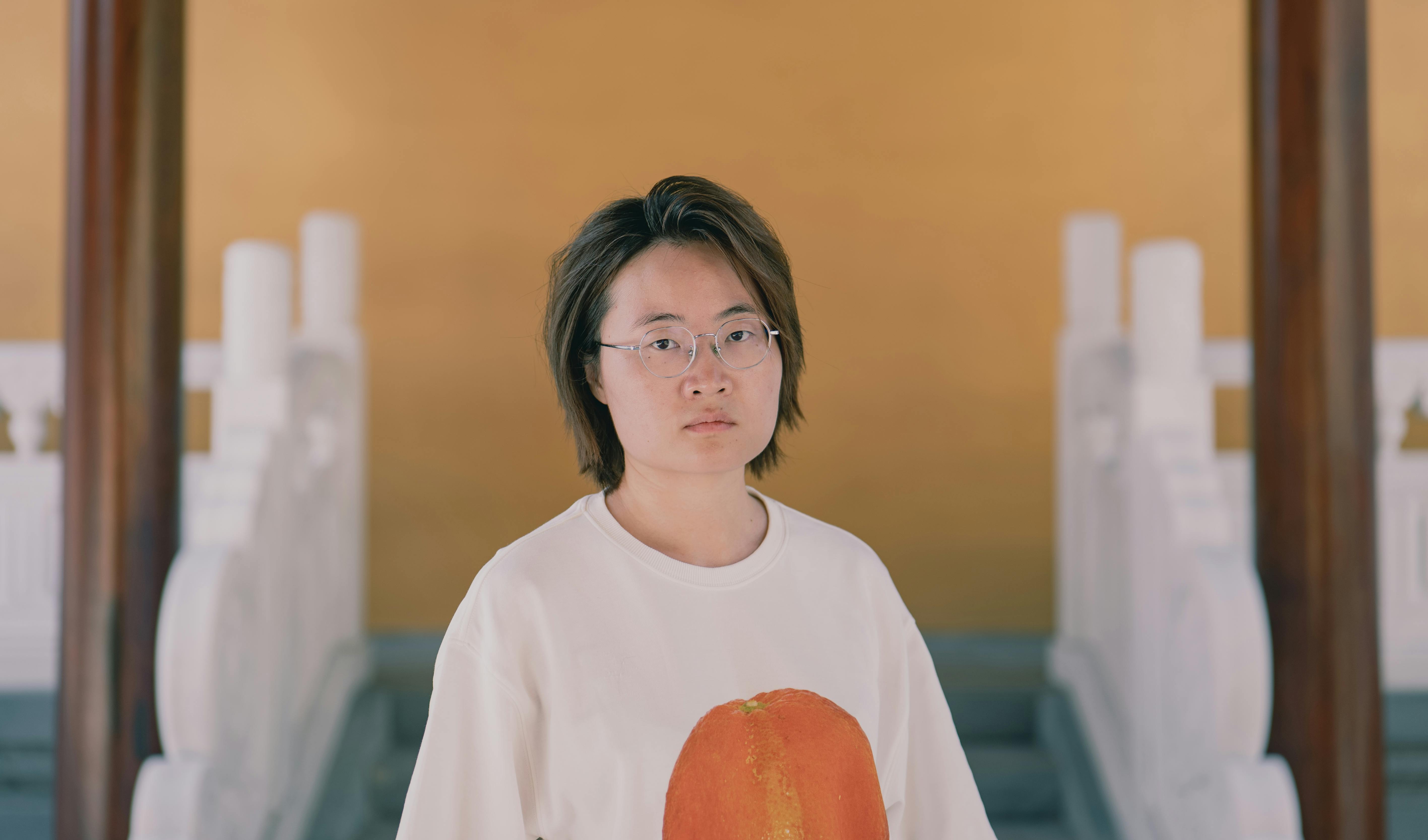 A young adult with glasses stands indoors wearing a casual t-shirt.