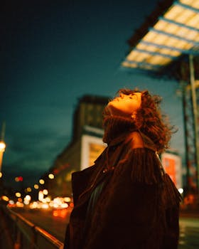 A woman in a coat looks up on vibrant city street during twilight.