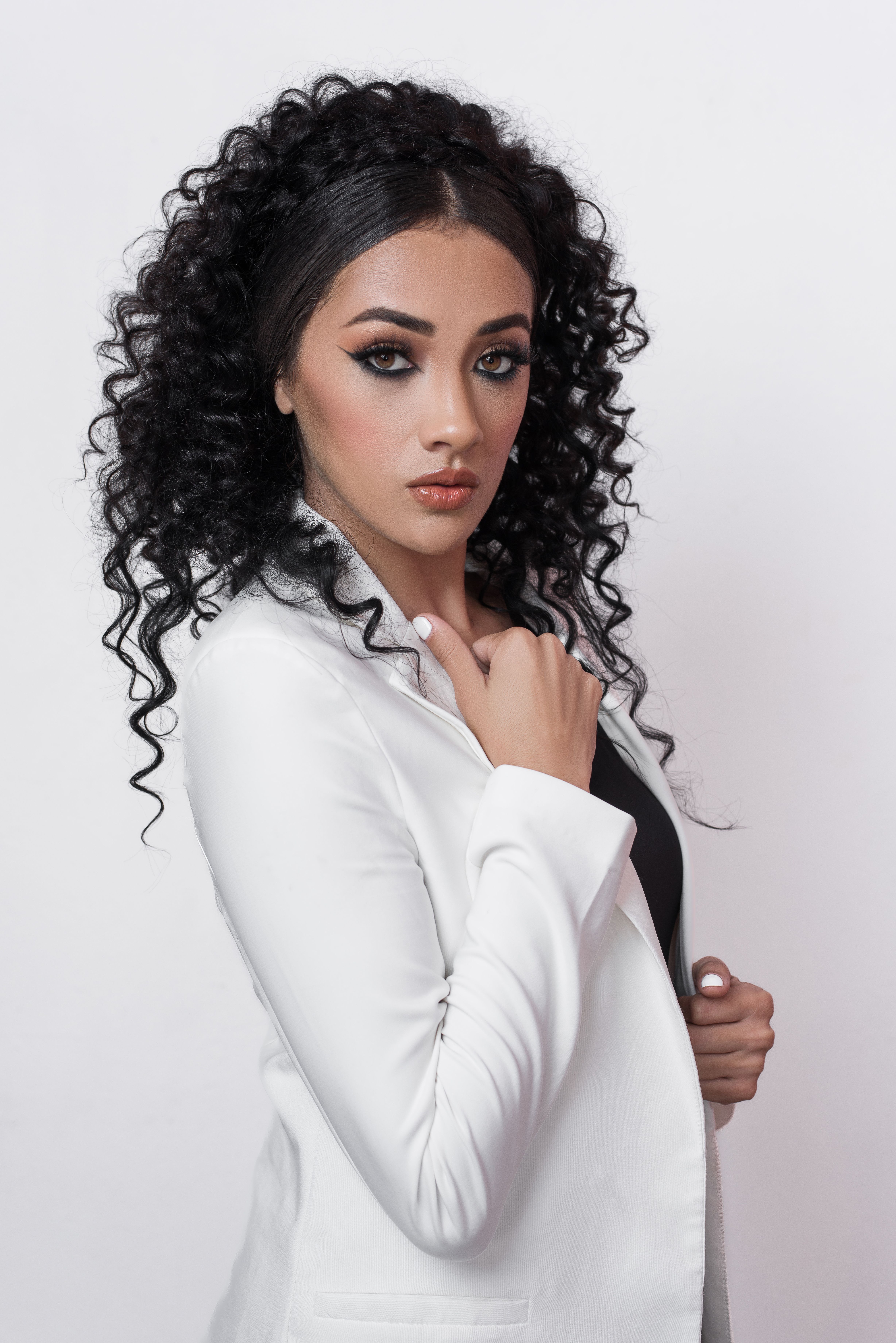 Stylish portrait of a brunette woman with curly hair in a white jacket posing confidently in a studio setting.