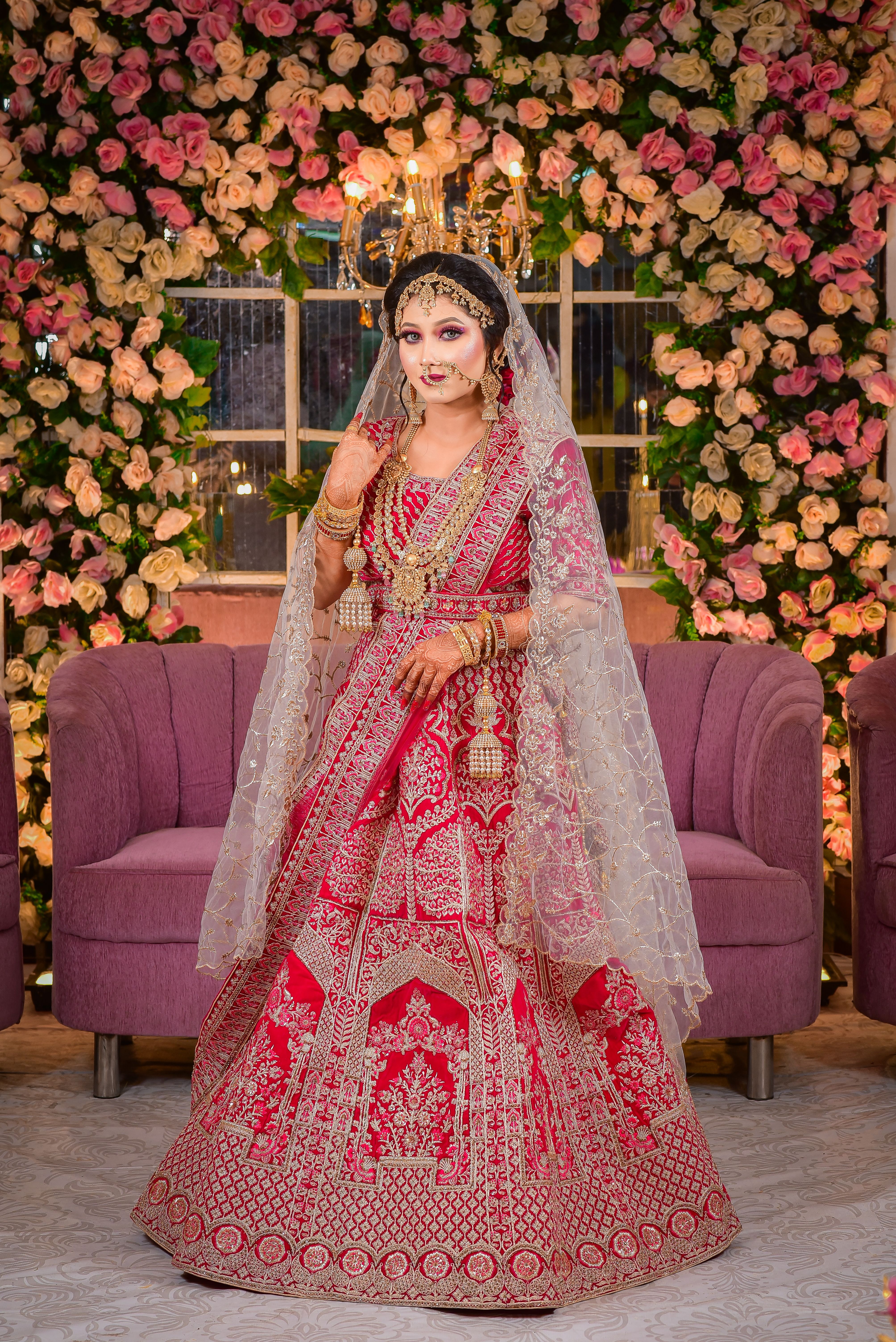 A South Asian bride poses in a red wedding dress with intricate jewelry against a floral backdrop.