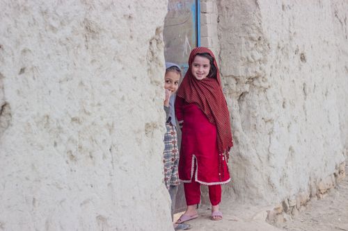Free Two girls in colorful traditional dresses smiling while standing beside a textured stone wall. Stock Photo