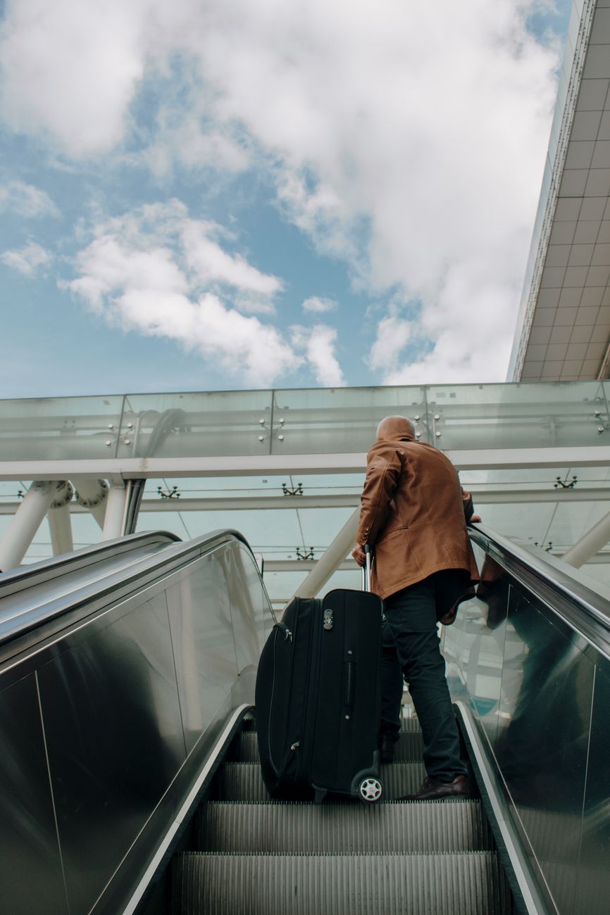 Volwassen man die met bagage op een roltrap in een luchthaven reist, met een heldere lucht boven zich.
