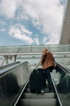 Adult male riding an escalator with luggage in an airport, traveling with bright sky overhead.