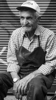 Black and white photo of an elderly man wearing an apron sitting on city streets.