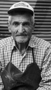Close-up portrait of an elderly man in a checkered shirt and cap, captured in black and white.