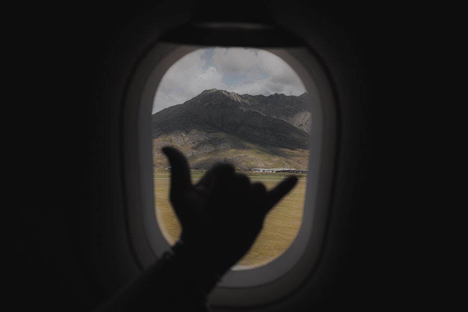 Hand making shaka gesture through airplane window with scenic Queenstown mountains in view.