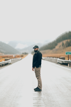 Bearded man in casual wear stands alone on a remote wet road, blending fashion with nature.