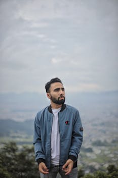 A bearded man wearing a blue jacket poses in a picturesque countryside with mountains in the background.