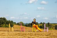A stylish woman wearing sunglasses sits on a colorful chair in a rural landscape under a bright sky.