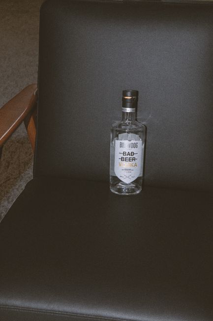 A bottle of clear vodka standing next to a pile of wheat grains and raw potatoes, showing two of the most common base ingredients used to distill it.