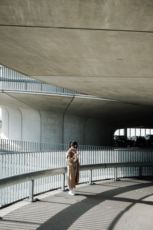 Free Woman in a trench coat standing in a modern urban setting with curved architecture. Stock Photo